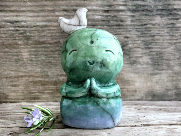 on a background of grey-brown driftwood, a raku ceramic jizo sculpture with a white bird on his head. he's is glazed is a bottom-to-top gradient of blue, forest green, and green, and he has a sweet smiling face and two cute praying hands. there's a sprig of flowering rosemary next to him on the left. 