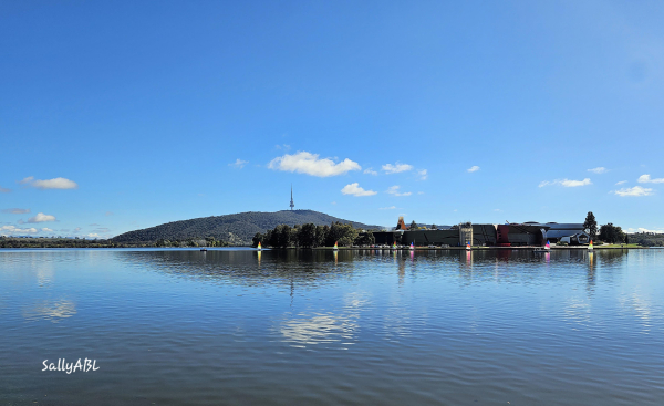 Photo taken April 2023 on a clear sunny Autumn day. Image shows wide shot of a lake with small colourful sailboats in the distance. On the horizon behindcthrm is a low mountain range with a tower at the top. A couple of small whispy clouds hang in a bright blue sky and are reflected on the water. The image  is calm, serene and quiet. 