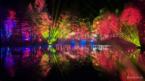 Photo of the view over the larger part of the loch, There are tall trees lit up in red, blues, greens and pinks, and they are reflected almost perfectly in the water on the bottom half of the photo.