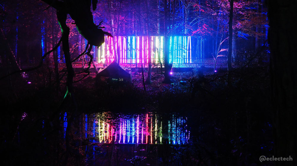 A photo of the tunnel from across the loch. From this angle it looks like a set of rainbow coloured vertical stripes with lit up trees behind and water in front. The water reflects the lights.