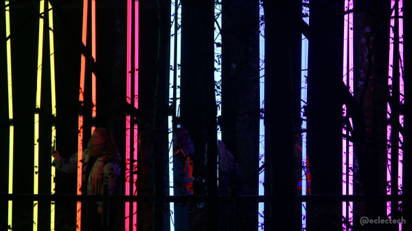 Photo of the tunnel from over the water, but zoomed in quite tight. The vertical strips of colour are bright, but they have jagged black areas cut out - the leaves and branches from surrounding trees. You can see people walking through the tunnel, in the darkness between the stripes.