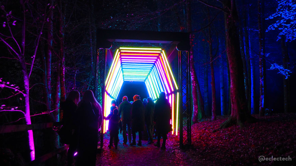 A photo of the tunnel from a few metres away. There is a group of people walking through, heads lit up reflecting the different colours lights from the angular archway. To the left and right you can see lit up trees in pink and blues.