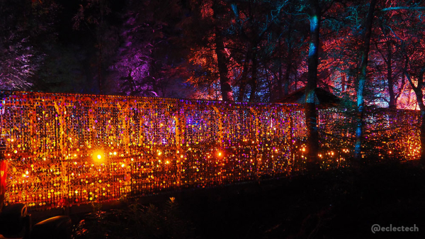 Photo of a wooden bridge across the loch. It has glitter ball style mirrored pieces dangling down the side, and is lit by orange lights. The trees behind are also lit in pinks and orange.