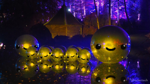 A photo of the large floating shiny balls lit up in yellow in front of a small wooden boathouse. I have drawn a smiling face on each of them,because of course I have. In the distance are tall trees lit up in blue and pink.