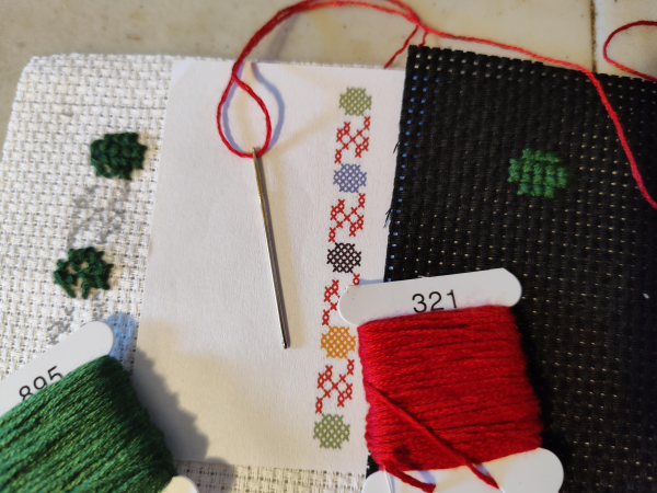 Close up photograph of some tatreez cross-stitching that is in progress. There are two small pieces of aida count 11 cloth, one white and one black. Between the two pieces of fabric is a tatreez pattern printed on paper of watermelons and vines. On top of the piece of paper rests a needle threaded with red thread.

The white cloth on the left has two green watermelons cross stitched in somewhat poorly. The rest of the pattern is drawn onto the fabric with pencil . Resting above this piece of cloth is a floss of green thread numbered 895.

The black cloth on the right has a single watermelon cross-stitched a little more evenly than the two on the white fabric. All three watermelons are green thread. Above the black cloth is a floss of red thread numbered 321.