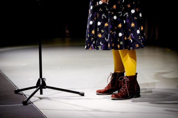 Photo from stage, only showing the bottom part of the microphone stand and my legs. I am wearing yellow tights, dark red lace-up boots and a black skirt with Halloween motifs all over it.