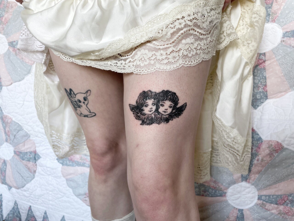 A black and grey tattoo of two cherub heads surrounded by wings on a thigh. There is another tattoo of a deer with butterfly wings on the other leg.