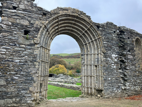 Photo of a rounded arched entrance doorway into the ruined Cistercian abbey of Strata Florida, the crumbled stone walls ending just above the arch. Through the doorway is a glimpse of the ruined interior walls, surrounded by  short grass, and of the hillside beyond. The leaves on trees growing in and around the abbey are turning green to gold