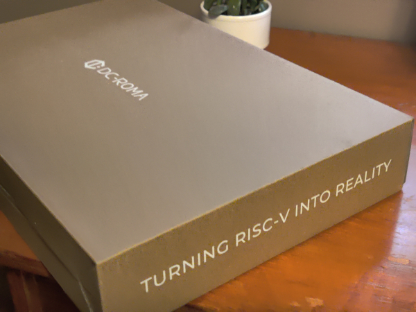 A cardboard box on top of a wooden desk. The front face of the box reads "DC-ROMA", and the side reads "Turning RISC-V into Reality".