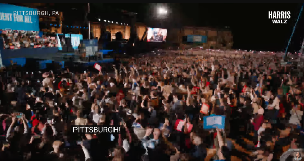 A large crowd of people are gathered outside at night. The crowd is cheering and waving their arms in the air. There is a large screen in the background with VP Harris on it. The texts "Pittsburgh" and "Harris Walz" are overlaid on the video image source.