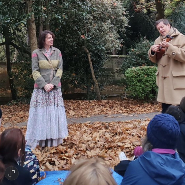 Vanessa and George standing in front of an audience seated on a tarpaulin in an autumnal park