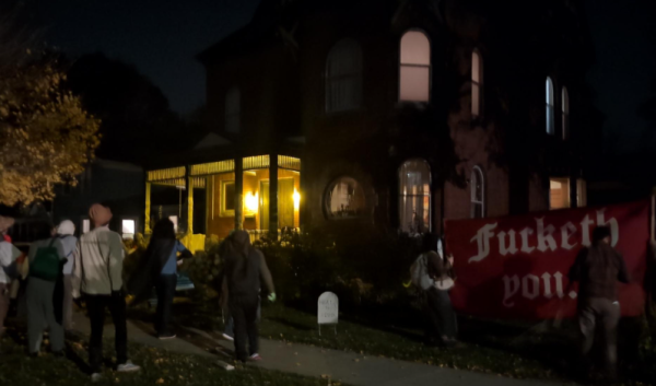 A crowd in various peasant attire stand in front of a large house in the dark. A prop gravestone is in the yard, next to a large banner reading "fucketh you" in a medieval font.