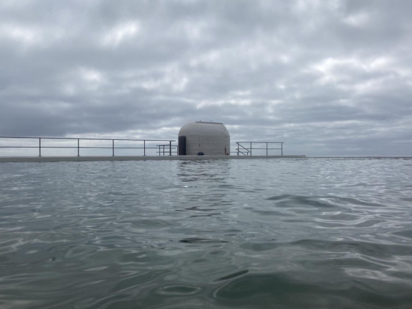 Photo taken while swimming, looking across the grey green water of the main pool to the rounded white pump house. Low clouds are pale grey with lighter highlights. 