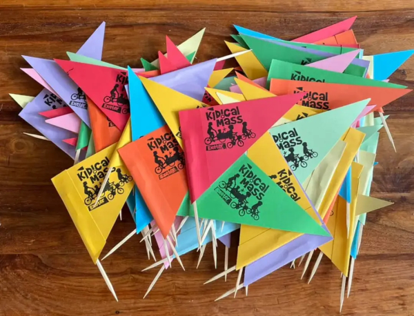 A pile of colourful bike flags screen printed with Kidical Mass Exeter and a logo of an adult on a cargo bike with a toddler and two small children cycling independently 