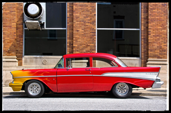 A picture of a 1957 Chevy Belair 2-door hardtop sedan with fins in the rear, parked at a curb in front of a brick building. The car, a real beauty, is painted like a flame, with yellow-gold in front turning to bright red by the time the paint reaches the driver's door.