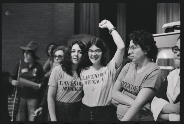 Black and white photo showing women wearing "Lavender Menace" shirts, with one lifting her fist up high and smiling at the camera