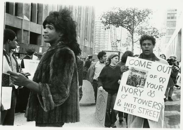 Black and white photo of a demonstration with Marsha P Johnson in the foreground and a person holding a sign saying "Come out of your ivory towers and into the street"