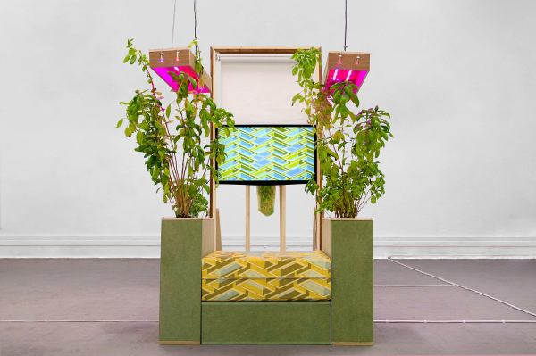 Sculpture of a hanging tv showing abstract animation, over a green and yellow geometric structure bookended by green plants beneath pink growing lights