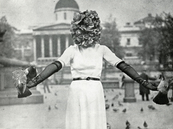 Black and white photo of a woman in a white gown and black gloves with her head obscured by a ball of roses, arms outstretched as pigeons land on her hands