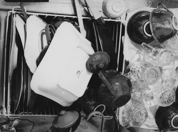 Black and white still of a kitchen counter with a drying rack loaded with plates, bowls, and glasses