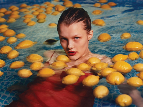 Photo of a young white woman wearing a red dress, floating in a pool filled with lemons