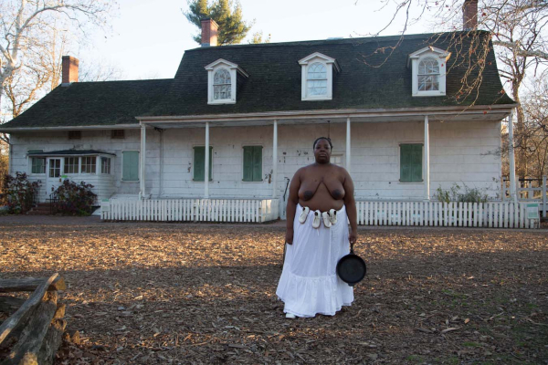 Photo of a Black woman in white heeled shoes, topless with a long white skirt, posing with a castiron pan in front of an old rural house on a leaf-strewn lawn