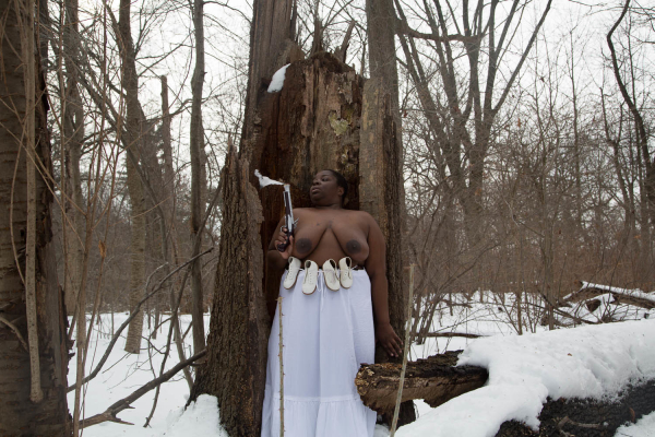 Photo of a Black woman in white heeled shoes, topless with a long white skirt, posing with a gun in a wooded winter landscape