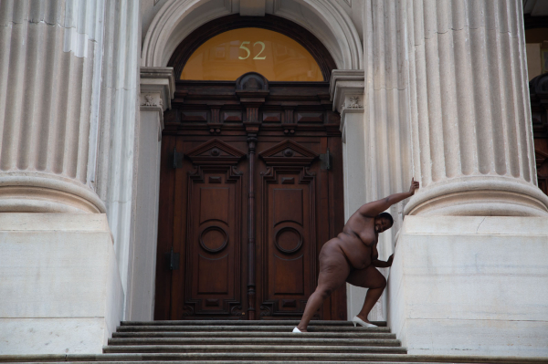 Photo of a nude Black woman in white heeled shoes standing in front of an imposing financial building, pushing her arms against its large white columns