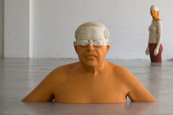 Carved wooden sculpture of a shirtless mustachioed man wearing goggles, seemingly erupting out of the floor like water. He is painted completely orange below his eyes.