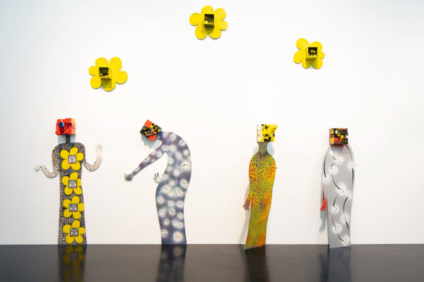 Four 2-D figurative sculptures with floral and other decorations posing against a white wall