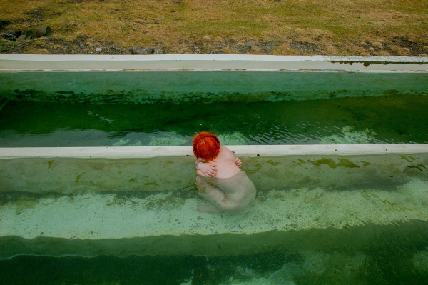 Photo of a white figure crouching hunched over in a green-tinted pool of water next to a brown-green field, with orange hair tied up in a braid, facing away from the viewer