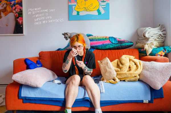 Photo of a white person with long orange hair in braids sitting on an orange couch surrounded by blankets, pillows, posters on the wall, and a hairless grey cat, holding up a syringe and filling it with testosterone. 