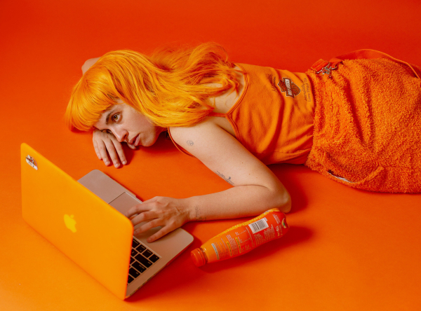 Photo of a white figure wearing an orange wig and orange top and skirt, lying down on an orange floor looking at a laptop with their hand on the mousepad