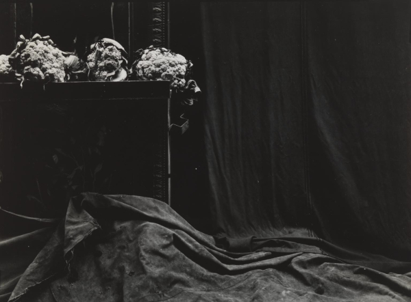 Black and white photo of an interior with a heavy wooden bureau with flowers on top surrounded by draping fabric