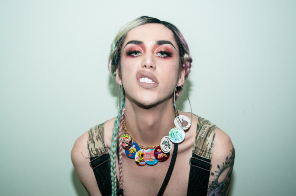 Photo of a drag performer looking directly at the camera with a sneering expression, against a white wall