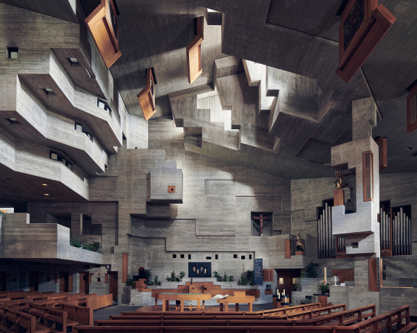 Photo of a Brutalist-style concrete church interior with wooden pews and multi-faceted sections throughout the walls and ceiling, and a skylight shining light down