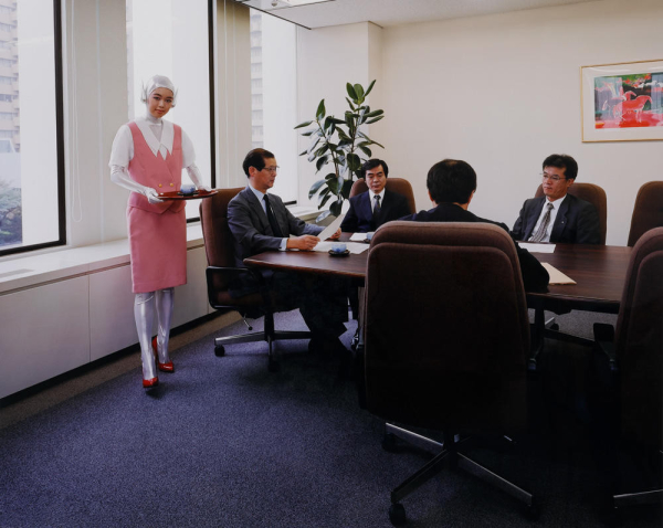 A young Japanese woman in a pink and silver outfit poses with a tea tray in a conference room where men in suits are sitting around a table