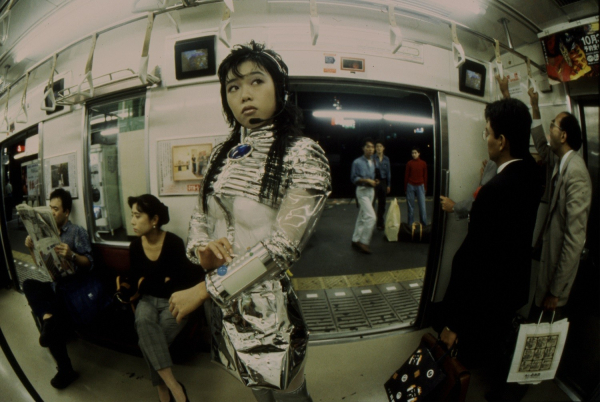 A young Japanese woman in a futuristic metallic outfit poses in the center of a subway train, with a fisheye lens effect.