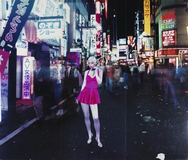 A young Japanese woman in a pink and silver outfit poses with a cell phone in a city street at night with bright signs and blurry figures all around her