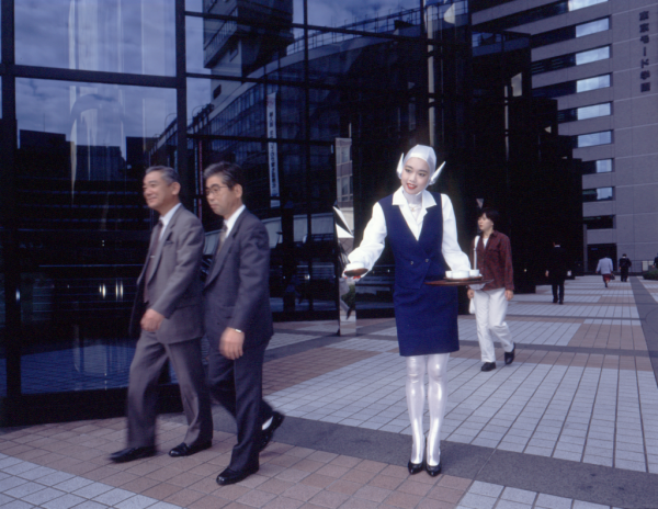 A young Japanese woman in a blue and silver outfit poses with a tea tray on a city sidewalk next to a glassy office building, with men in suits passing her by