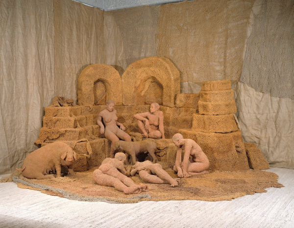 Installation of a group of light brown fiber sculptures of human figures and a pig lounging around stacked architectural forms