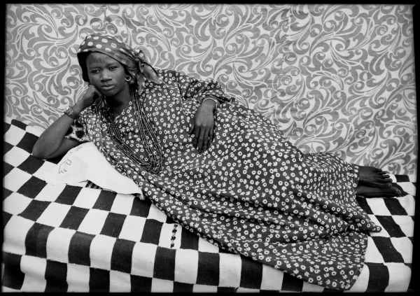 Black and white photo of a Black woman in a patterned dress and head scarf lounging on a bed with a checker pattern