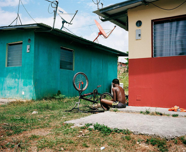 Photo of a young Black man seen from behind and afar, sitting between teal and red houses working on an upside down bike