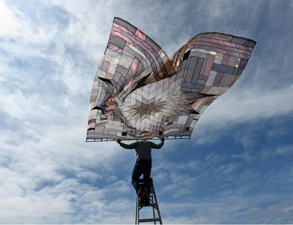 Photo of a figure standing on a ladder against a cloudy sky, holding a large quilt with a star pattern made with shiny and transparent fabrics, billowing in the wind above