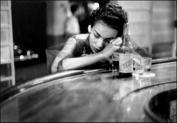 Black and white photo of a young Black woman leaning on a curved bar, her head resting on her hands in front of a glass bottle and several glasses of alcohol