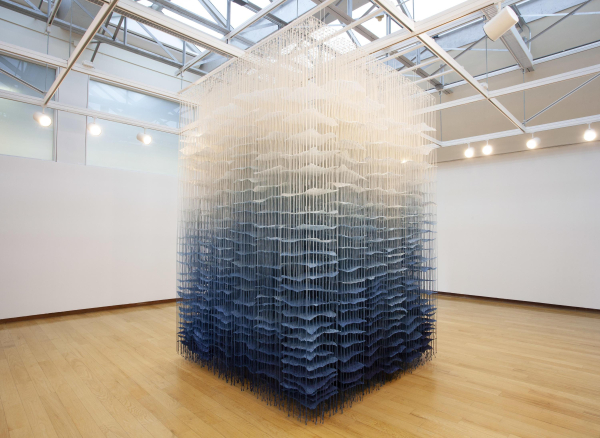 Hanging sculpture installed floor-to-ceiling in a gallery, made up of long cascading threads with strips of paper hanging within, arranged in vertical layers, dyed a blue-to-white gradient moving upwards