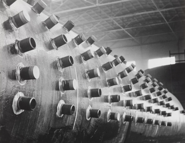 Black and white photo of a large, curving piece of metal stretching across the frame, covered in metal pipes jutting out in vertical rows