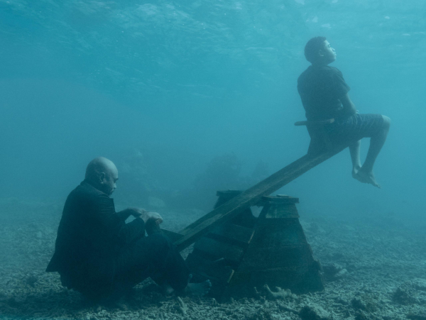 Underwater photo of a man in a suit sitting on the lower end of a seesaw, with a younger figure sitting on the upper end