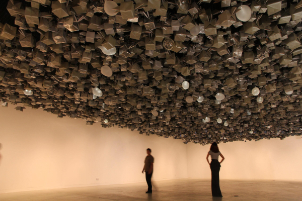 Installation of a large room with hundreds of metallic forms, both square and round, hanging from the ceiling in a jumbled cluster