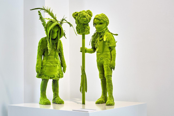 Sculpture of two childlike figures standing together, one holding a staff with a broccoli-like top, all entirely in a bright green hue, with leafy forms around their head and neck